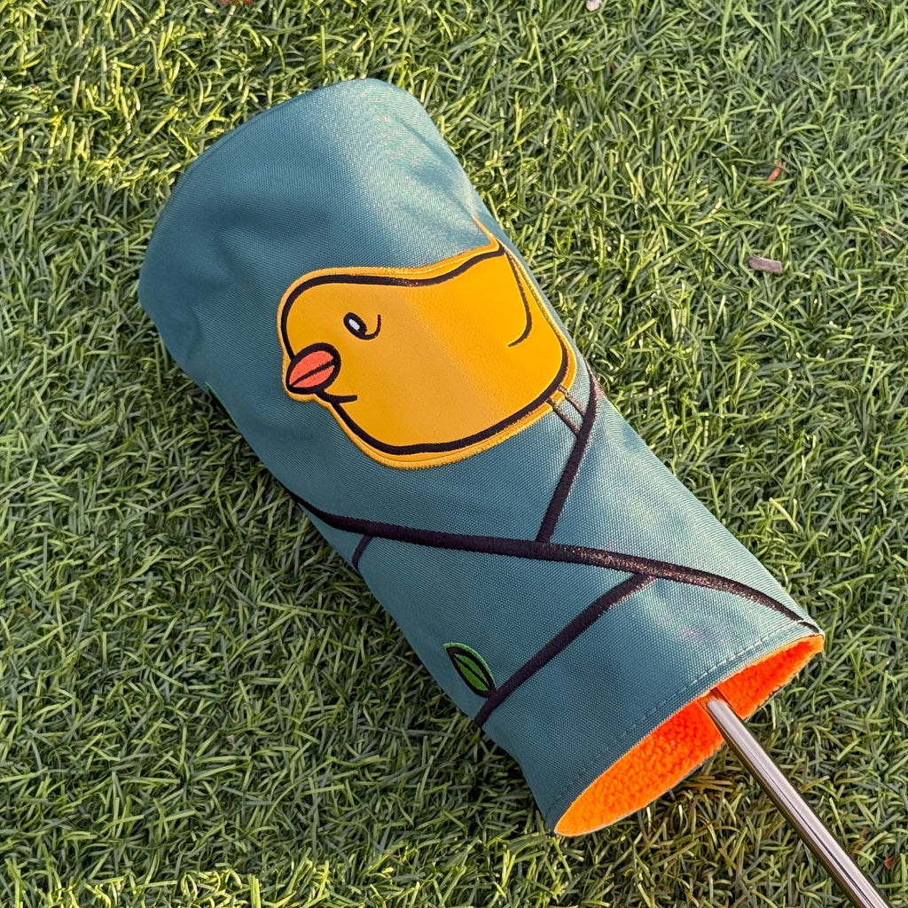 A golf club putter head cover lying on green grass, featuring a yellow cartoon sneak bird graphic on a muted blue-green background with an orange interior lining visible at the opening.